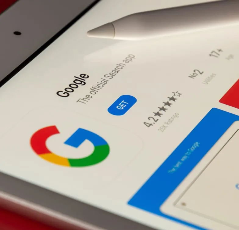 Focalizando la aplicación de Google en la pantalla de un iPad, magnificada por un lápiz óptico, se destaca por su altísima calificación por parte de los usuarios. Esta perspectiva cercana sirve para valorar la importancia que Google le da al SEO para mantener su elevada reputación. Con el objetivo de dominar el mercado del SEO, Google ha optimizado su aplicación no sólo para ser amigable con los motores de búsqueda, sino también para satisfacer las demandas y necesidades del usuario final. Resultando en una puntuación de usuarios elevada en todas las tiendas de aplicaciones y plataformas móviles.