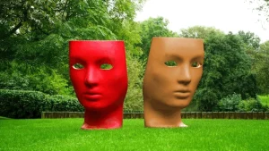 Dos colosales cabezas esculpidas que brillan en tonos rojos y beige, estratégicamente ubicadas en medio del lujoso y verde césped, forman un sorprendente contraste con el fondo rústico adornado con árboles.