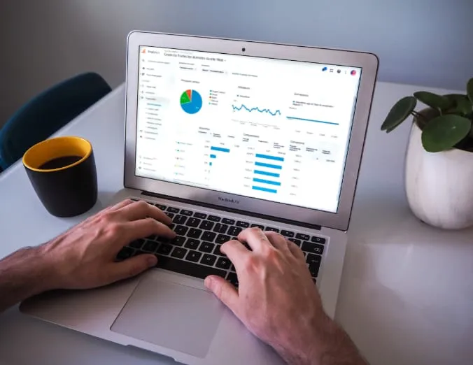 Un experto en análisis de datos está examinando gráficos estadísticos en la pantalla de su computadora portátil. Acompañándole en este escenario de trabajo, una taza de café y una planta verde decoran el escritorio.